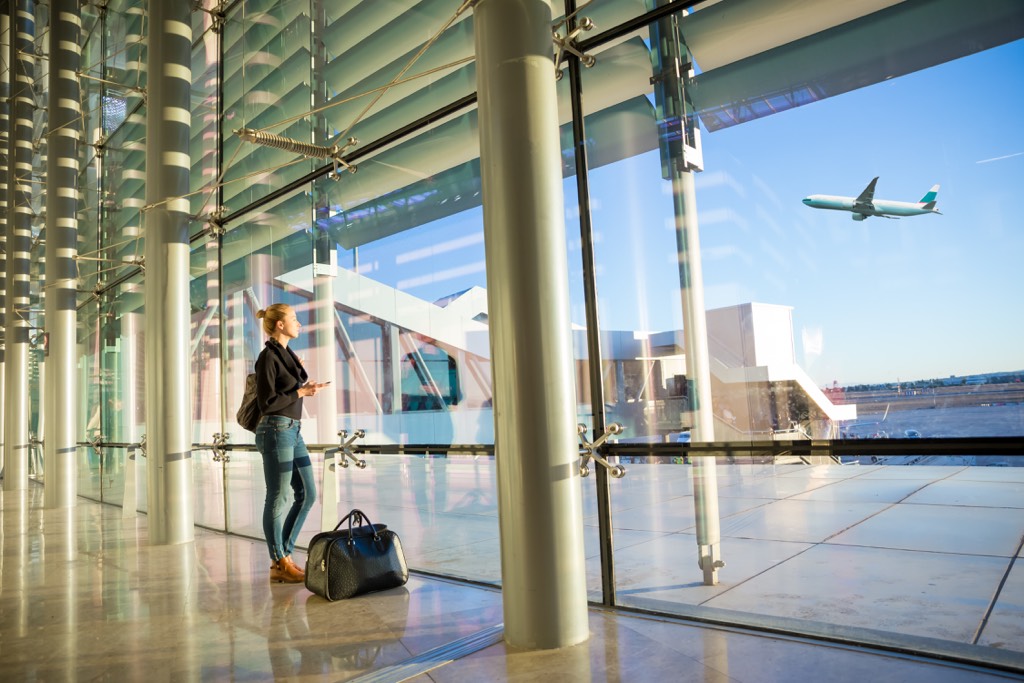 Young casual female traveler at airport, holding smart phone device, looking through the airport gate windows at planes on airport runway.
